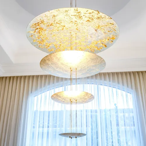 A multi-tiered chandelier with four glowing circular discs, the top one gold-textured, against an arched window with sheer curtains.