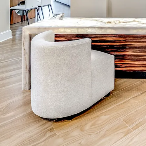 A cream-colored curved boucle chair on a light wooden floor, next to an illuminated marble counter with a dark wood-patterned base.