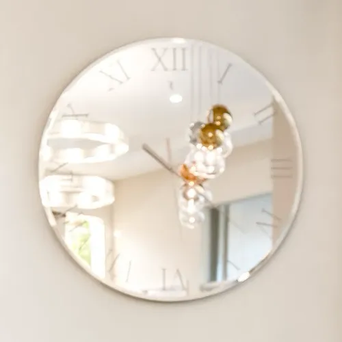 A round, frameless mirror clock with Roman numerals reflecting a modern spherical chandelier and interior space.