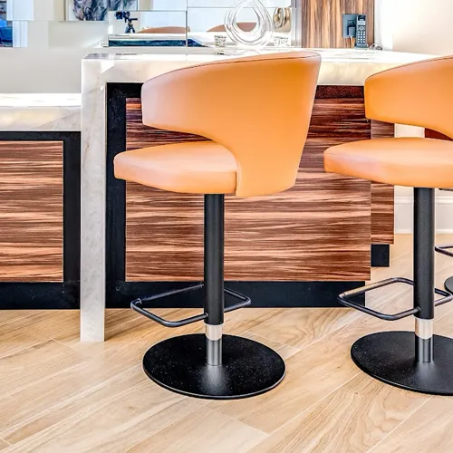 Two tan leather bar stools with black bases in front of a kitchen island with a white marble top and dark wood cabinetry.