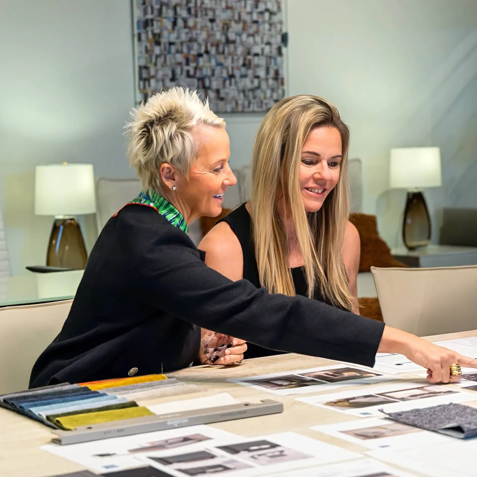 Two smiling women, one with long blonde hair and one with short spiky blonde hair, sit at a table, pointing at design documents and fabric swatches.