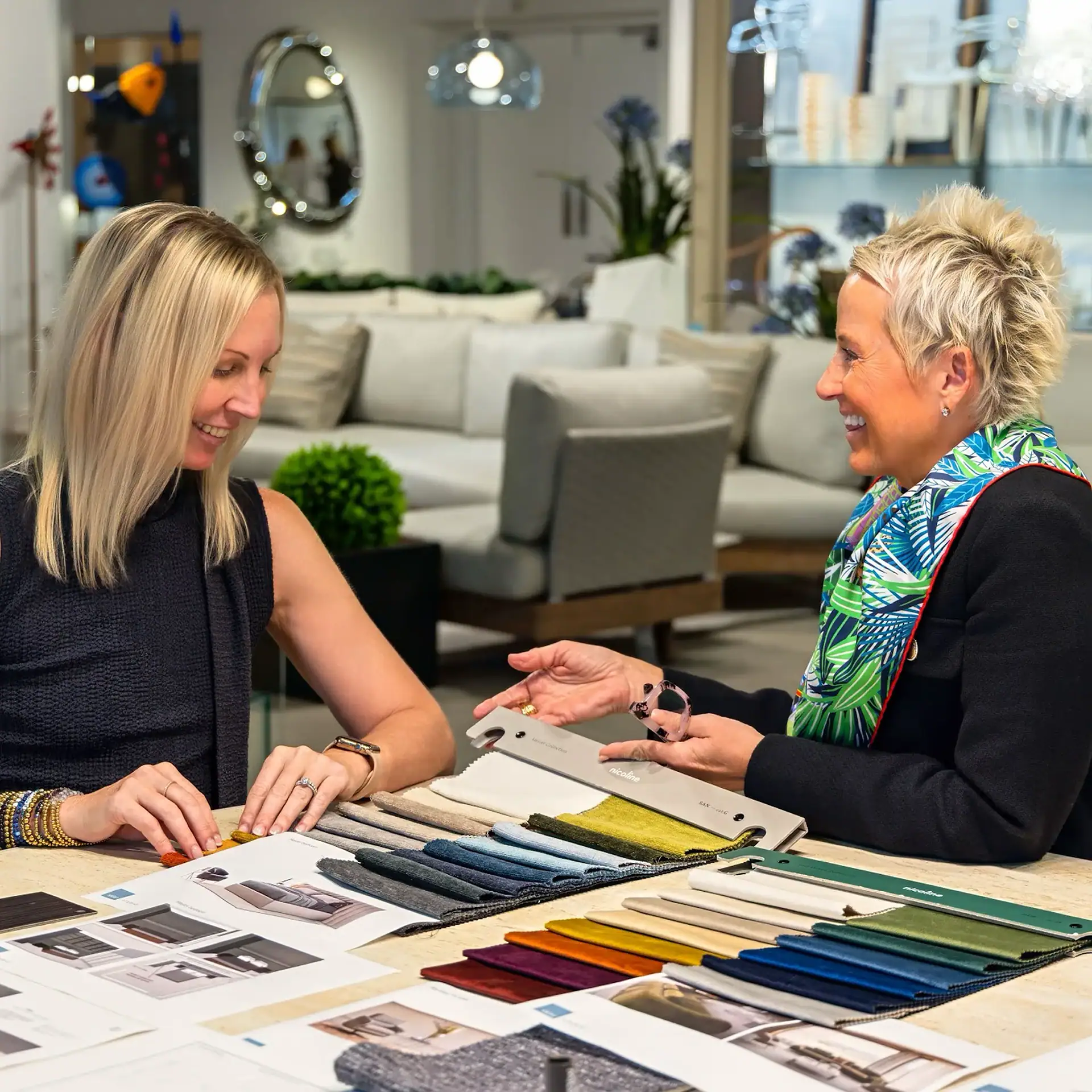 Two women smiling and discussing fabric samples and design documents at a table in a showroom.