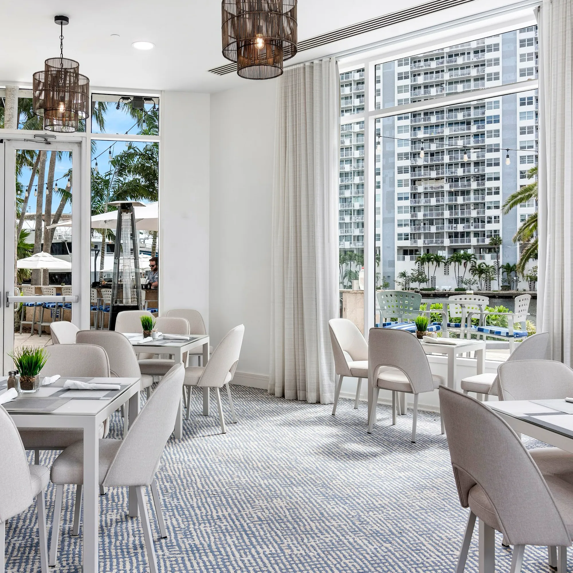 A modern dining room with white tables, beige chairs, and a blue and white patterned carpet, featuring large windows overlooking an outdoor patio with palm trees and a city skyline.