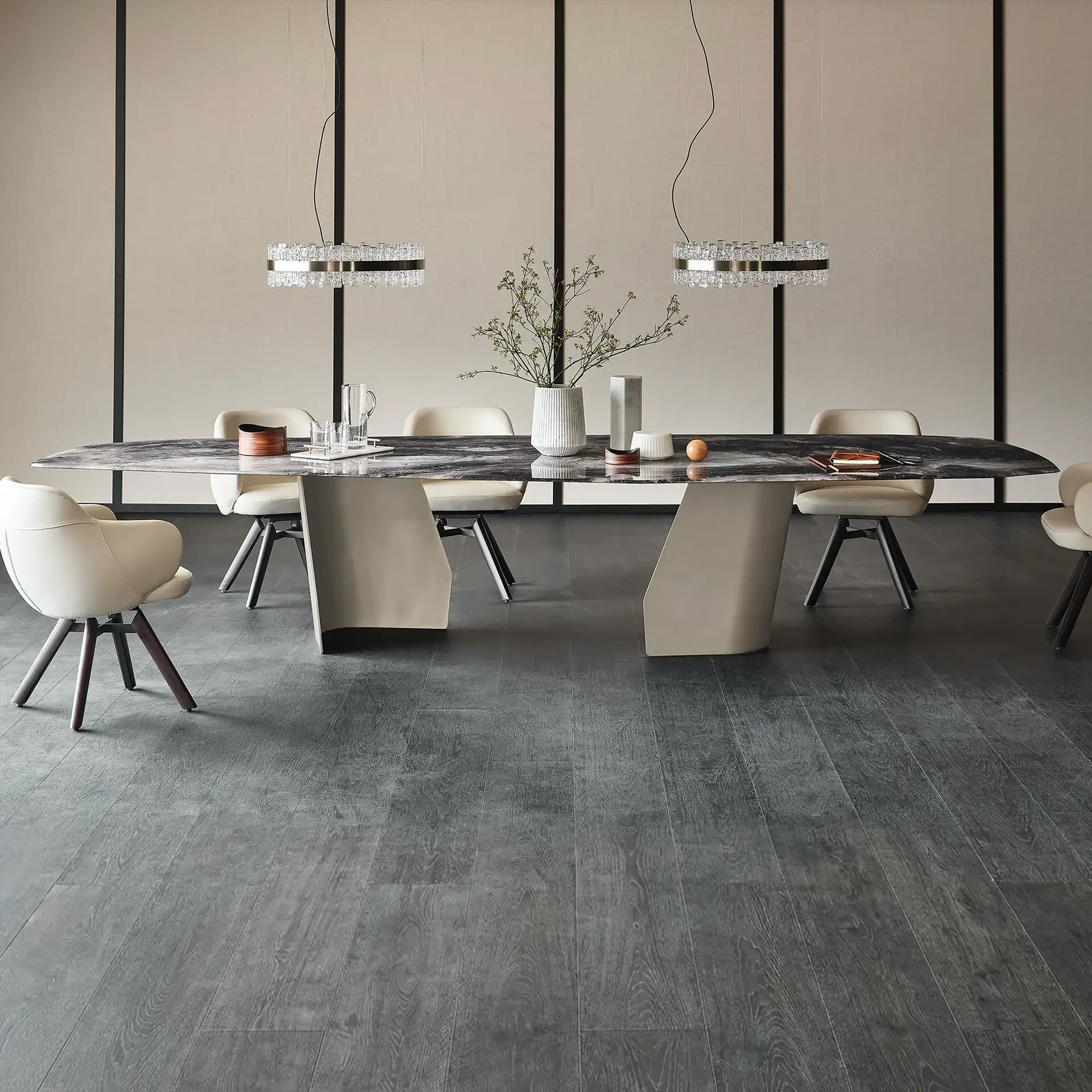 A modern dining room with a long marble table, six cream chairs, two crystal pendant lights, and a paneled beige wall.