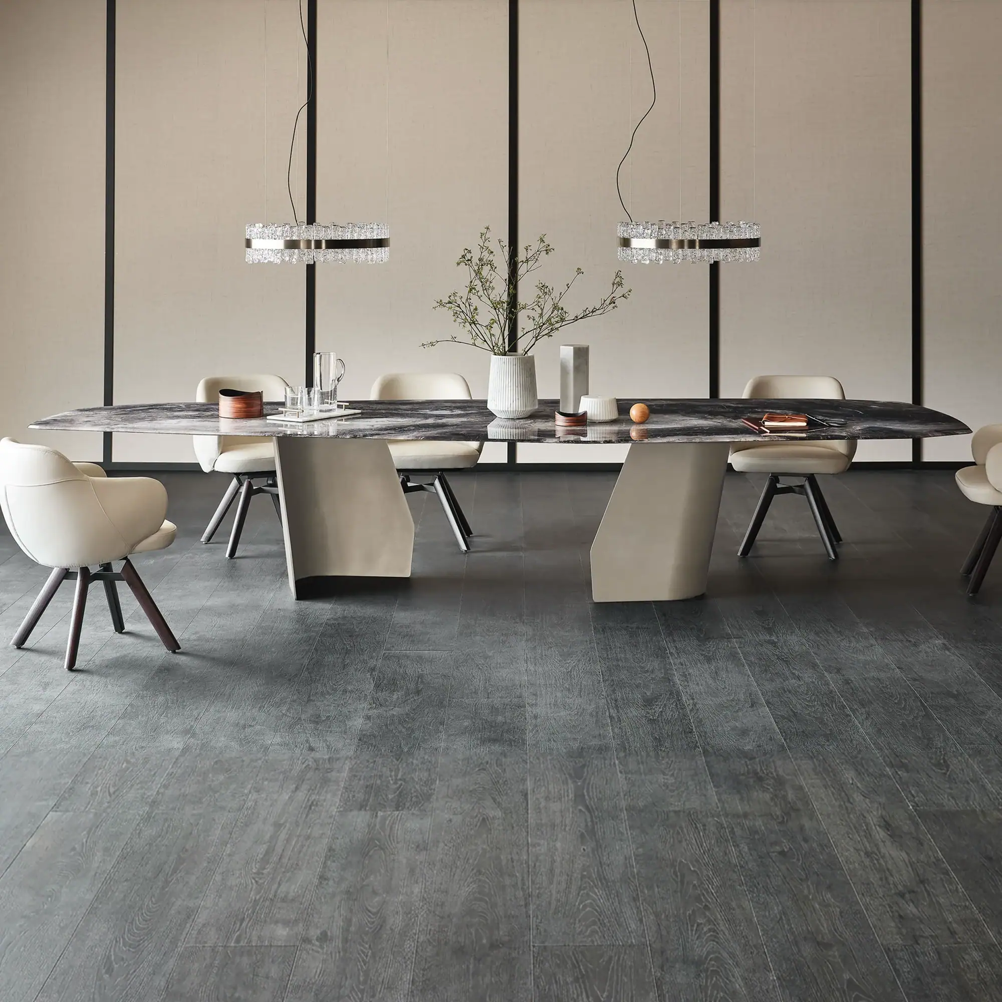 A modern dining room with a long marble table, six cream chairs, two crystal pendant lights, and a paneled beige wall.