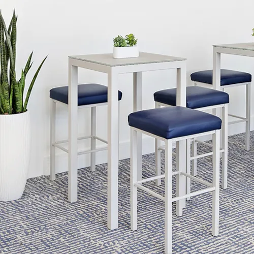 Two white bar-height tables with dark blue stools on a blue and white patterned carpet. A tall snake plant in a white pot is on the left.