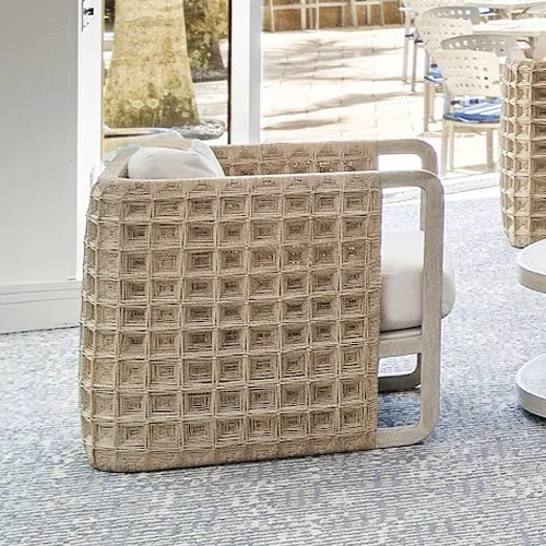 A modern outdoor armchair with a light-colored woven square pattern and light cushions, set on a patterned rug with an outdoor patio and palm trees in the background.