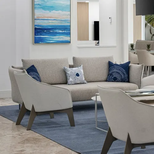 A modern lounge area with a beige sofa, blue decorative pillows, light-colored armchairs, a blue rug, and a large seascape painting on a white wall.
