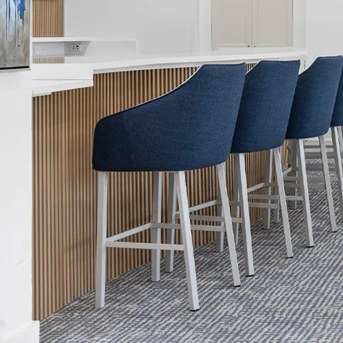 Four navy blue bar stools with light frames in front of a white counter with vertical wooden slat paneling. The floor has a blue and gray patterned carpet.