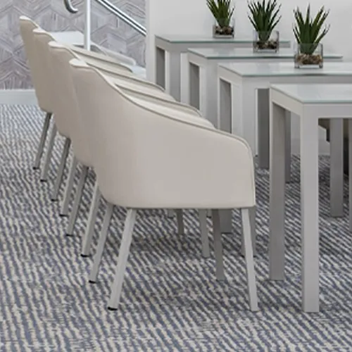 A row of light beige chairs with gray legs on a blue and white patterned carpet, next to three gray tables with glass tops and small green plants.