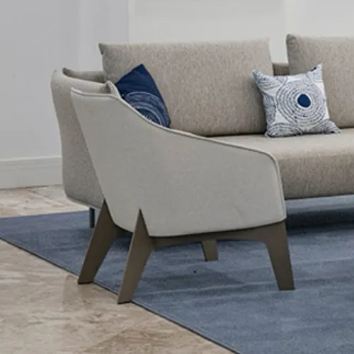 A light gray armchair with dark legs in front of a beige sofa with blue and white patterned pillows, all on a blue rug.
