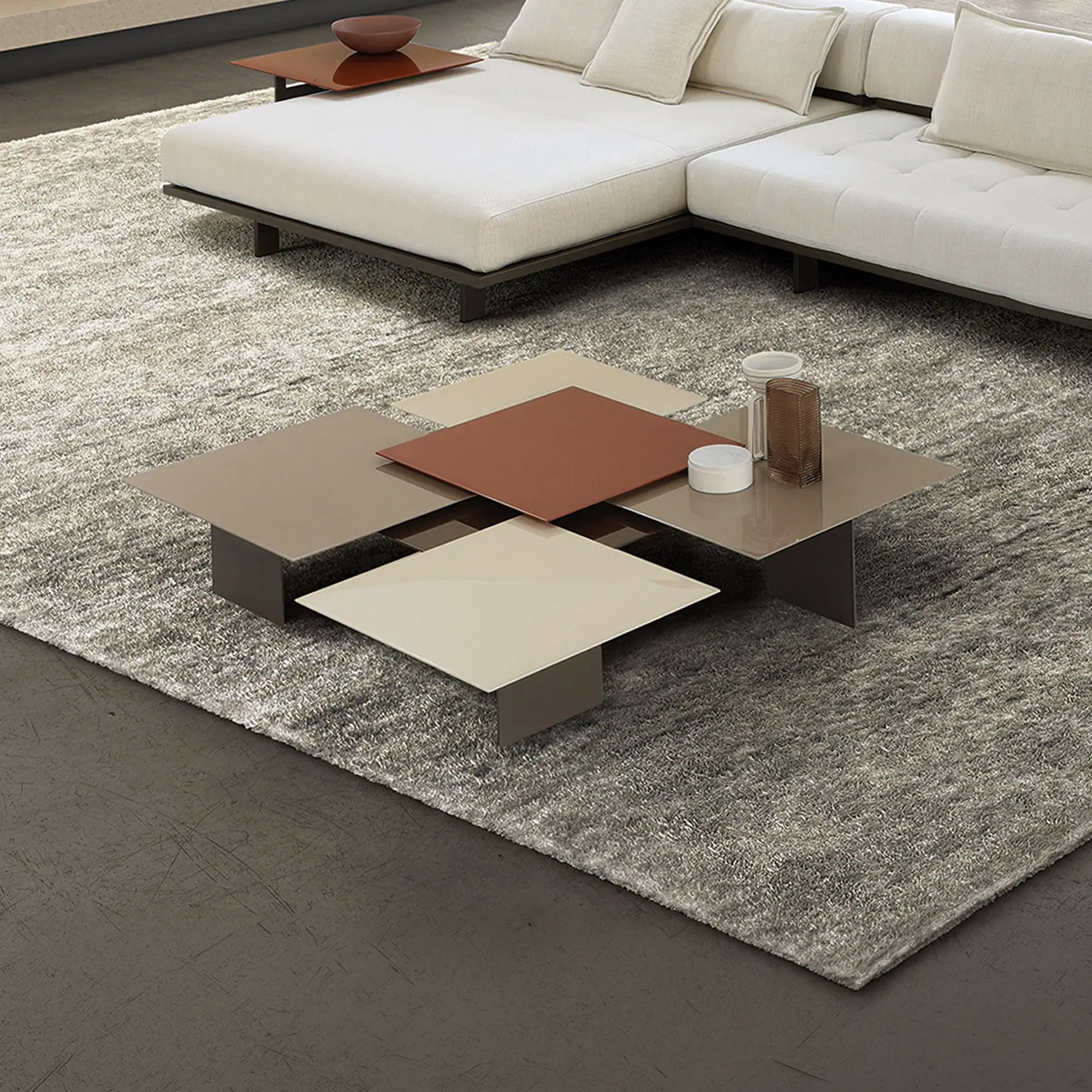 A modern living room featuring a cream L-shaped sectional sofa, a unique modular coffee table with brown and beige surfaces, a textured grey rug, and a fireplace with a warm fire. Large windows in the background overlook green trees.