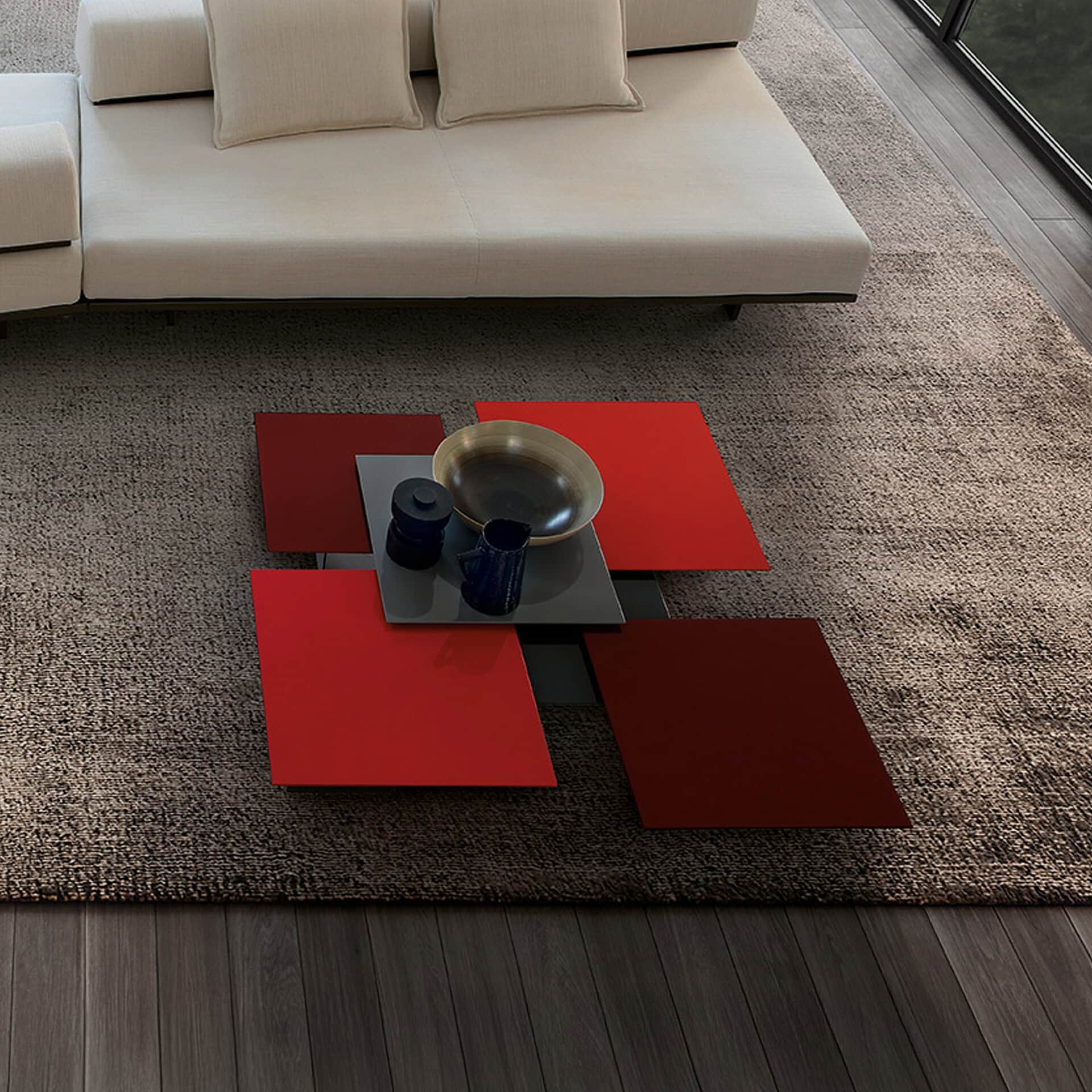 A modern living room featuring a unique modular coffee table with bright red and dark burgundy square panels, a light beige sofa, and a textured grey rug. Decorative items are on the table.