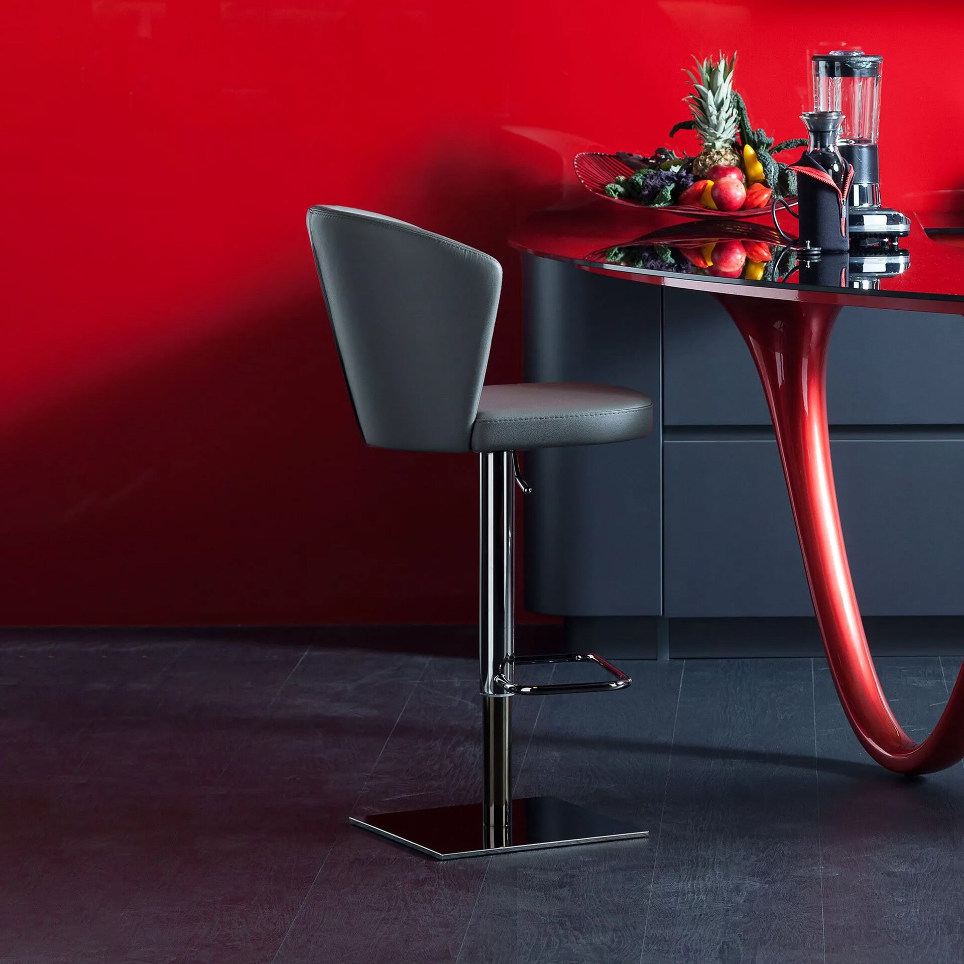 A modern kitchen scene with a gray bar stool, a red and black island with a glossy black top and curved red base, and a fruit bowl with a blender, set against a vibrant red wall.