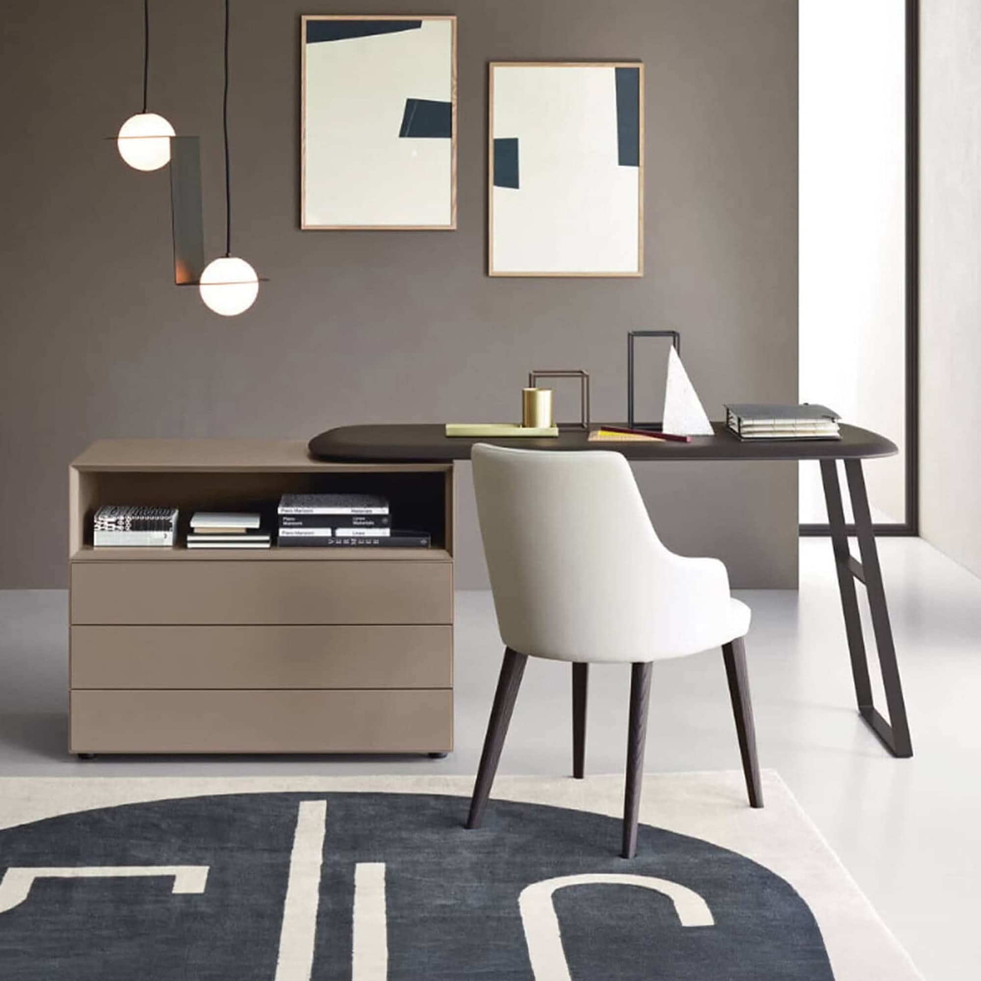 A modern home office featuring a dark brown desk, integrated beige storage unit, and a white upholstered chair. The dark gray wall is adorned with abstract art and two spherical pendant lights. A dark blue and white abstract rug covers part of the light-colored floor.
