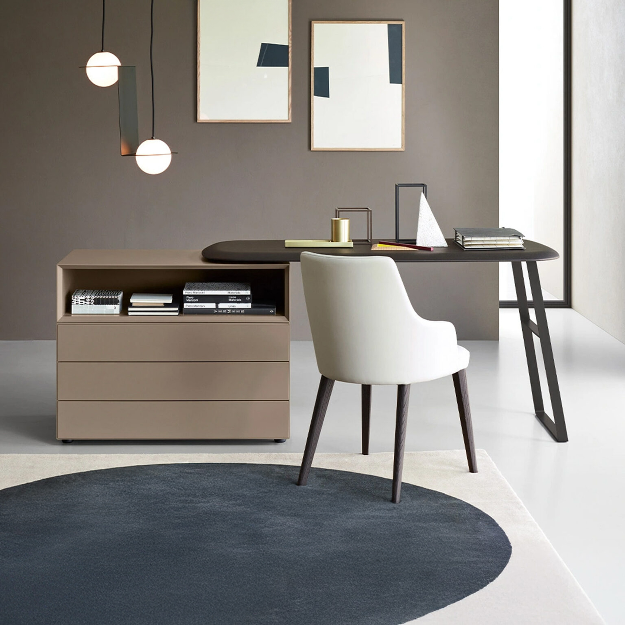 A modern home office featuring a dark brown desk, integrated beige storage unit, and a white upholstered chair. The dark gray wall is adorned with abstract art and two spherical pendant lights. A dark blue and white abstract rug covers part of the light-colored floor.