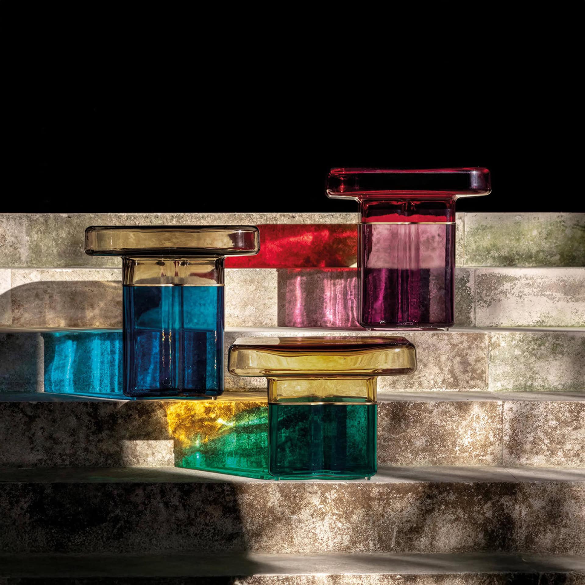 Three modern glass objects in blue, purple, and green with golden tops, arranged on textured grey steps. Strong light creates colorful reflections on the steps.