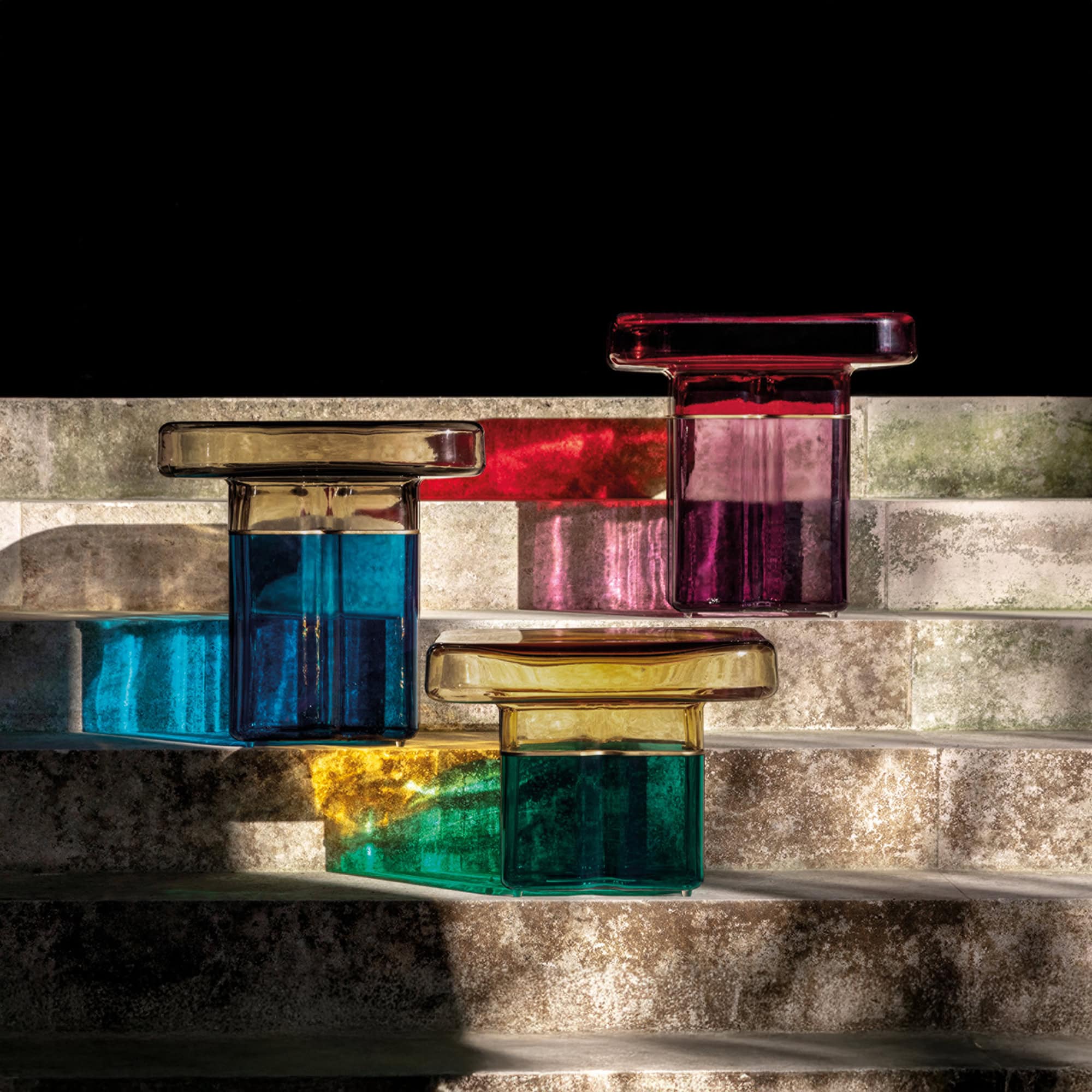 Three modern glass objects in blue, purple, and green with golden tops, arranged on textured grey steps. Strong light creates colorful reflections on the steps.