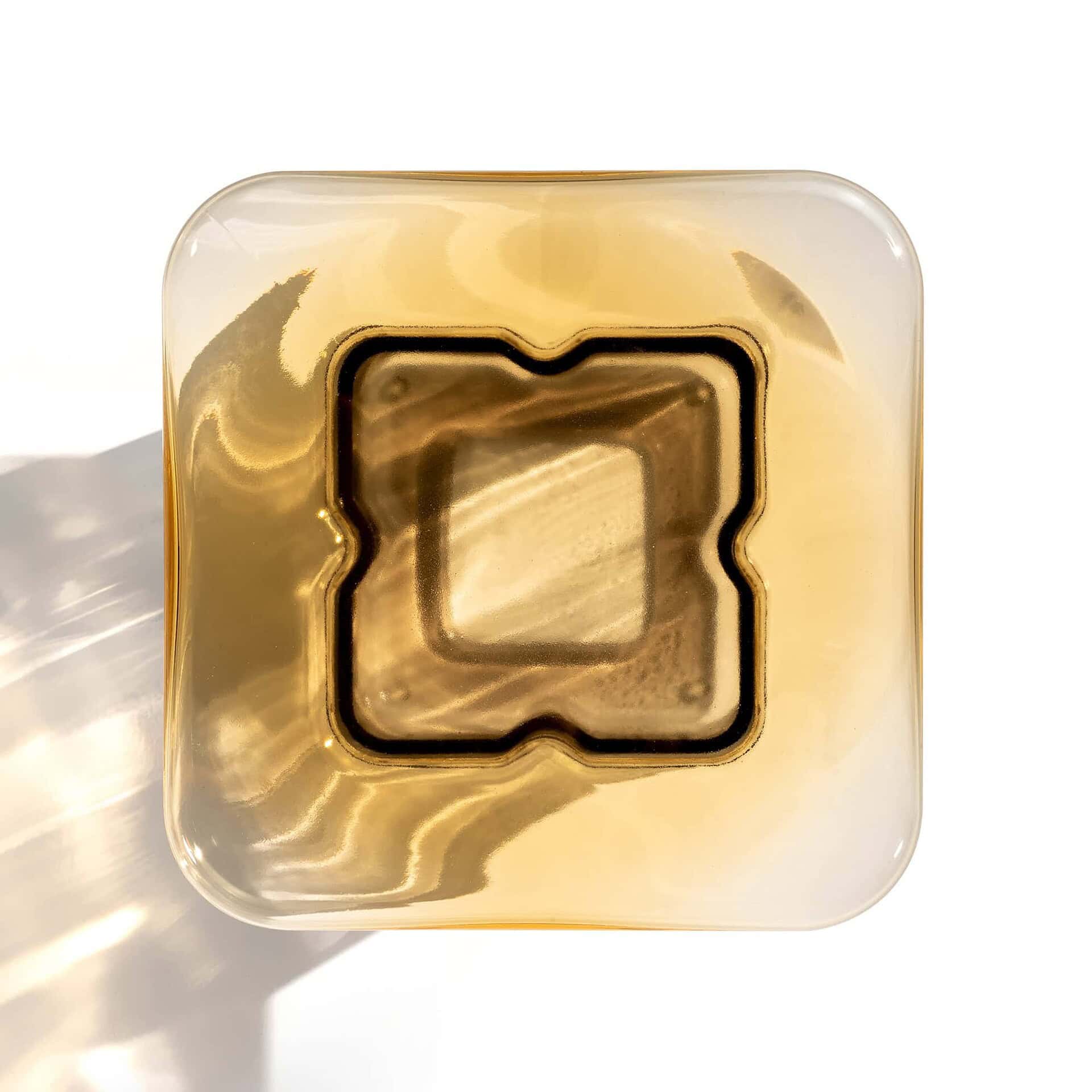 A square, amber-colored glass or resin object with rounded corners and a recessed, stylized square pattern in its center, set on a white background with light reflections.