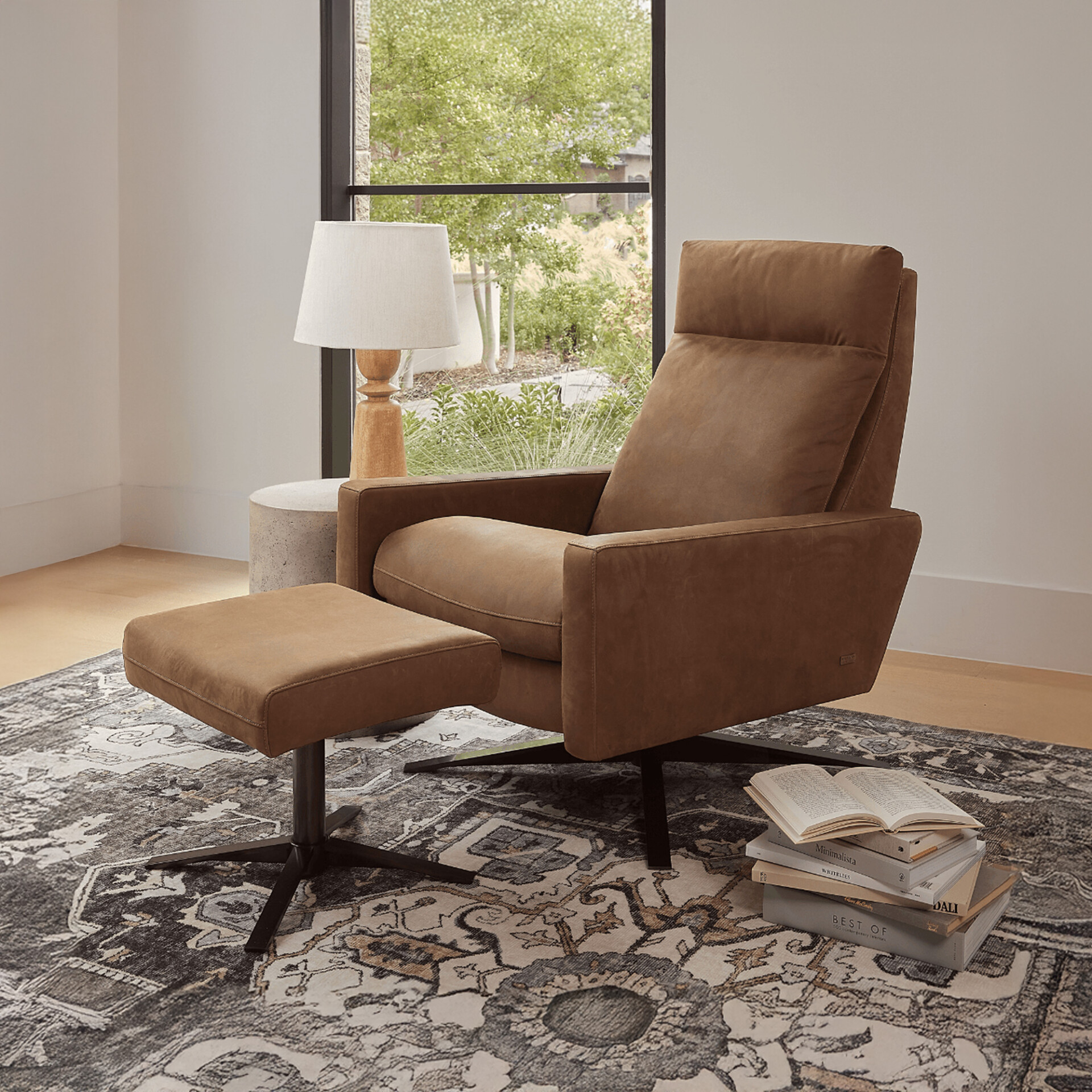 A modern brown leather recliner and matching ottoman in a bright room with a large window overlooking green trees. A patterned rug, a side table with a lamp, and a stack of books complete the cozy scene.