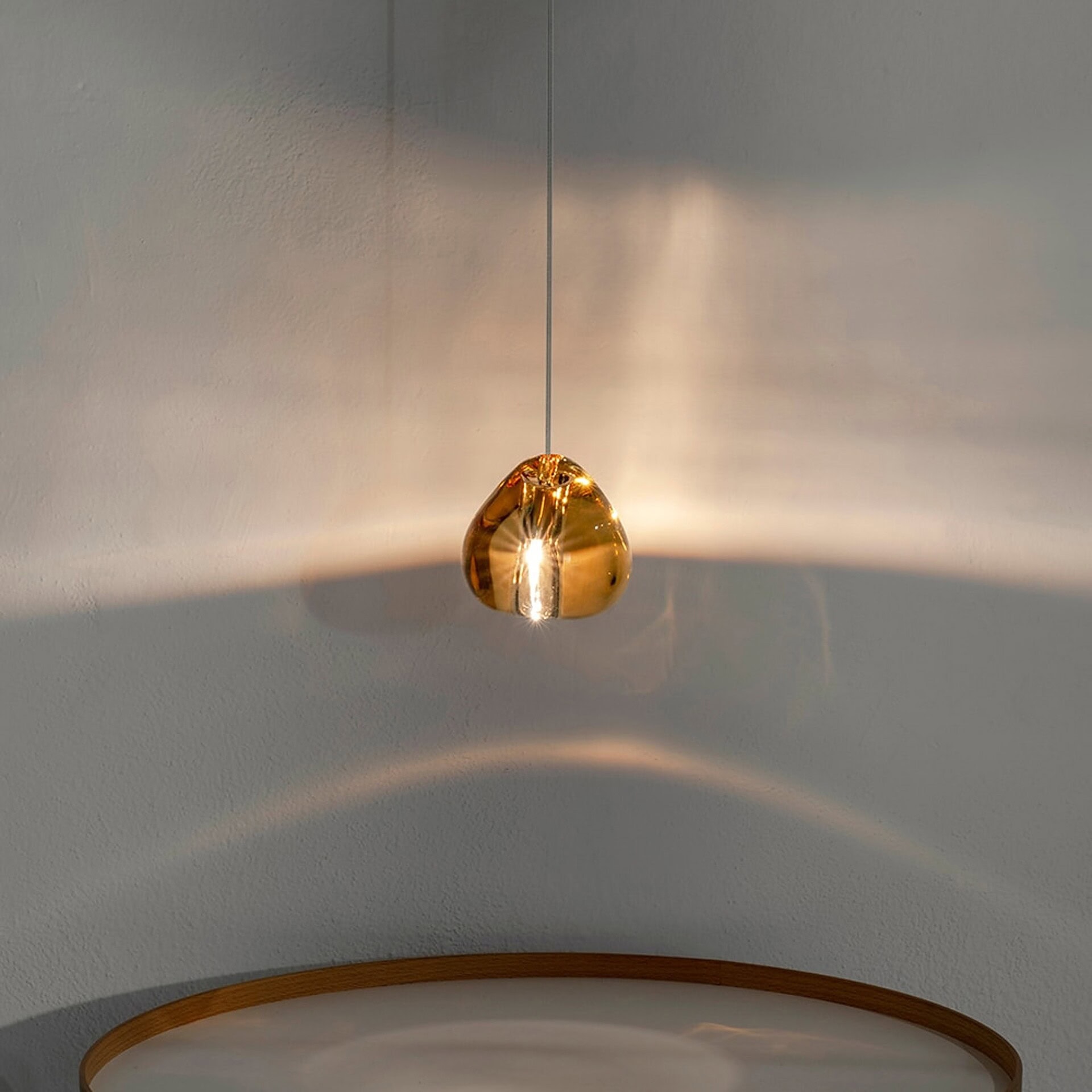 A modern, organic-shaped pendant light made of reflective amber-gold glass hangs against a textured light wall, casting warm light and reflections. A wooden-rimmed table is visible below.