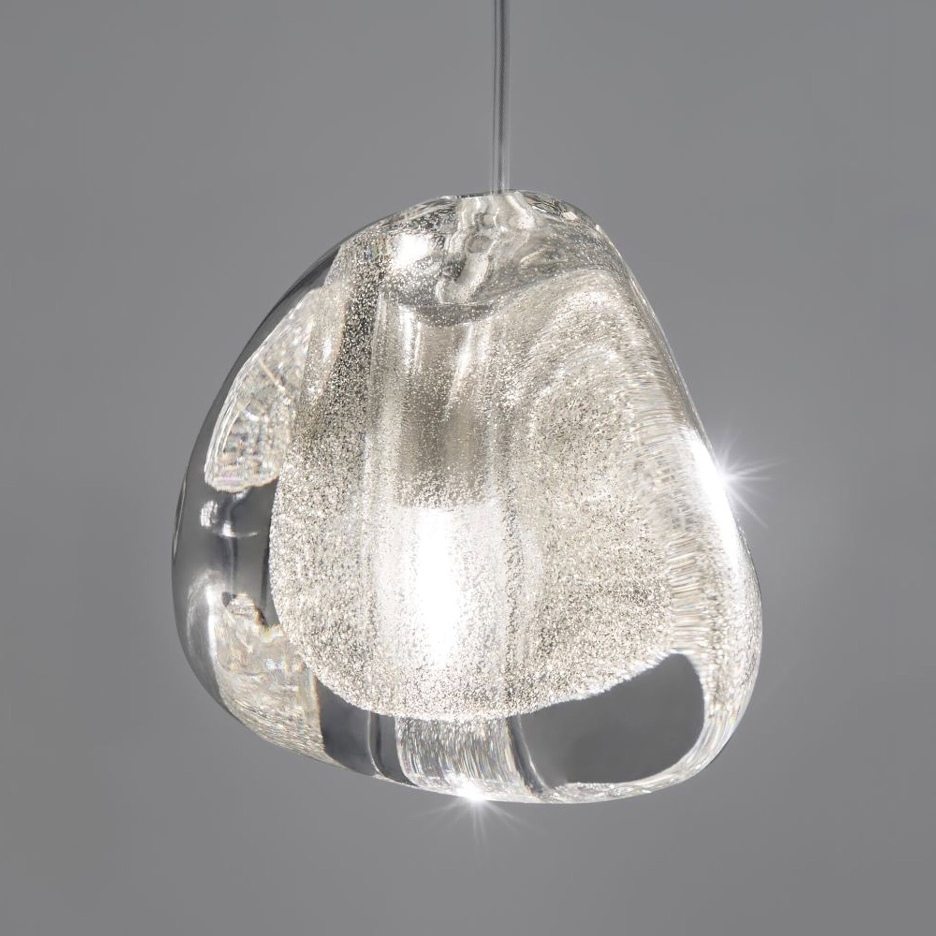 A modern, clear glass pendant light with an irregular, organic shape, featuring a sparkling, bubbly interior and a bright internal light source, suspended against a gray background.