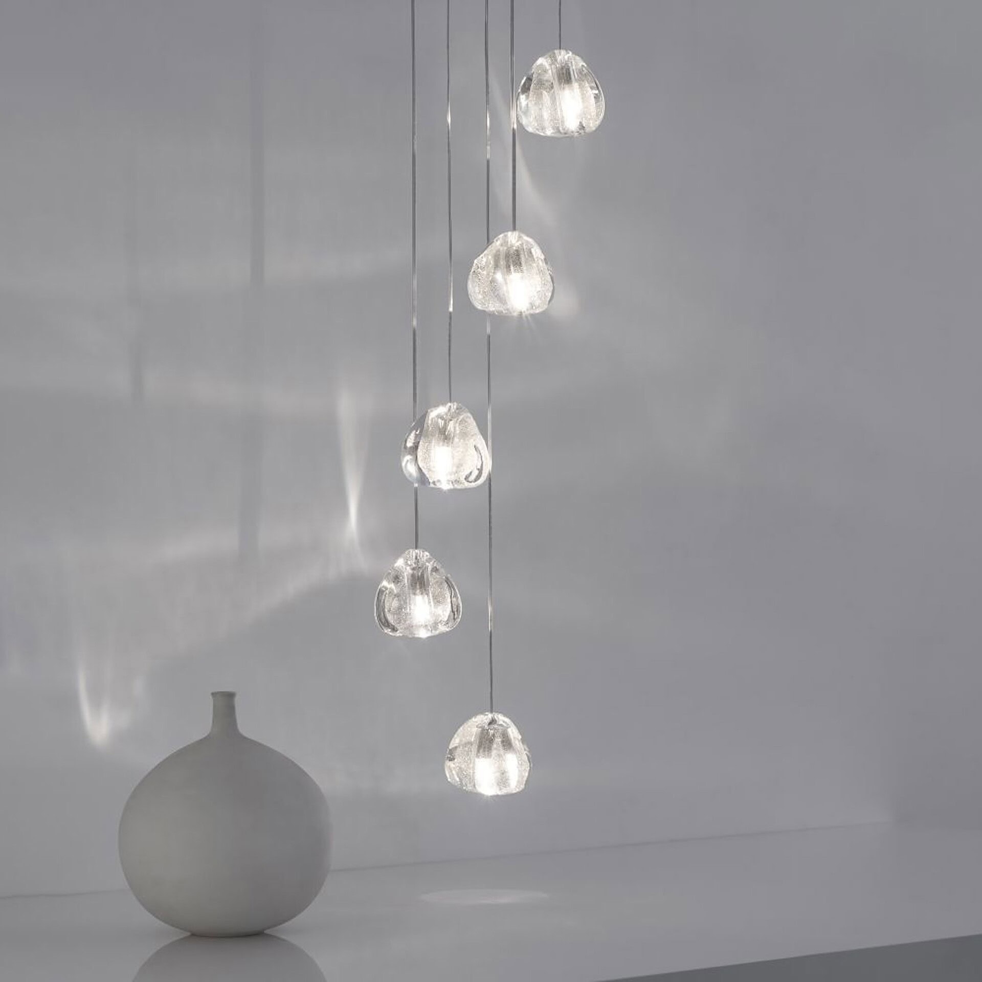 Six illuminated, textured clear glass pendant lights hang at varying heights against a light gray wall, with a matte white ceramic vase on a reflective surface below.