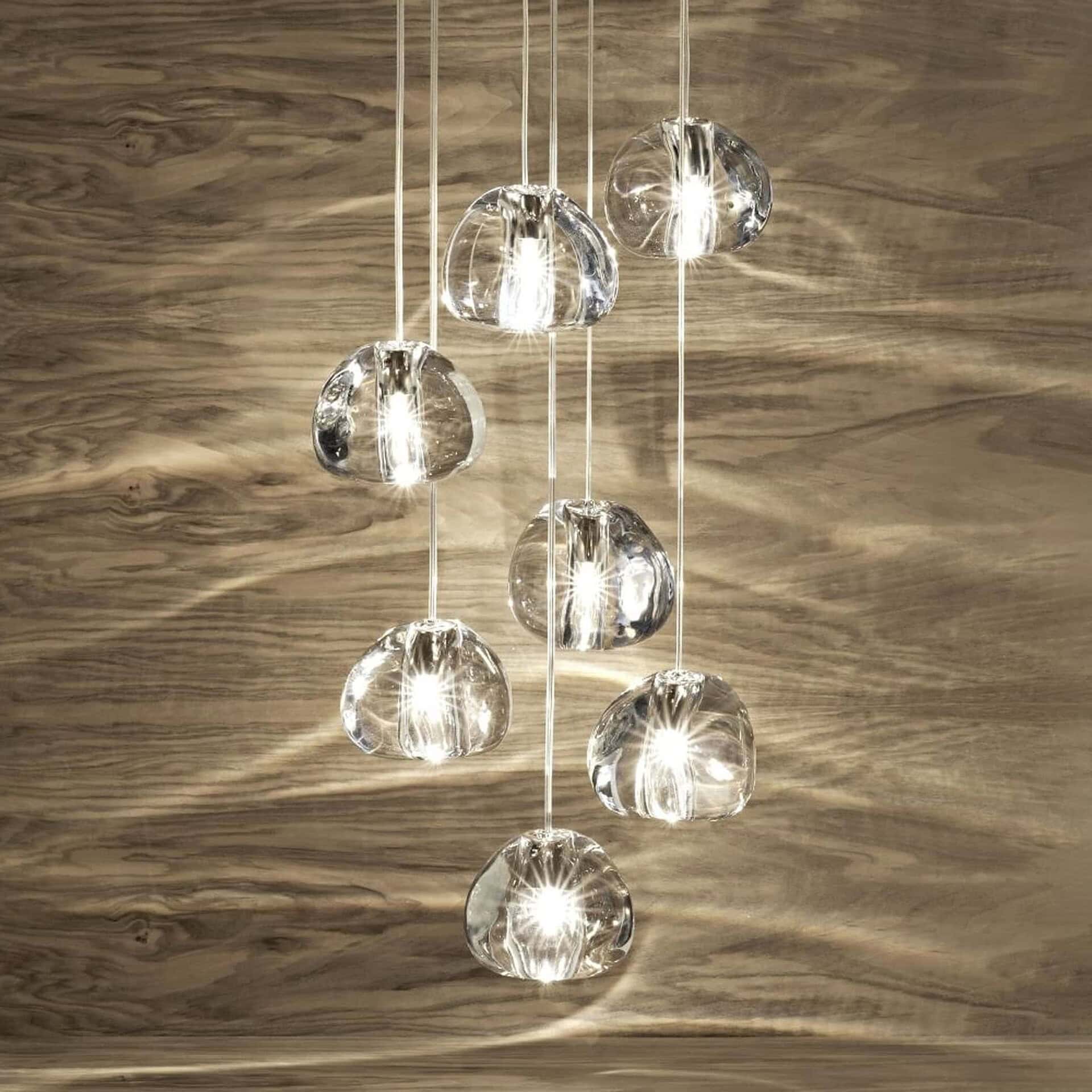 A multi-light pendant fixture with eight clear, irregularly shaped glass globes suspended at various heights against a warm, horizontally grained wooden wall. The lights are illuminated, casting bright reflections.