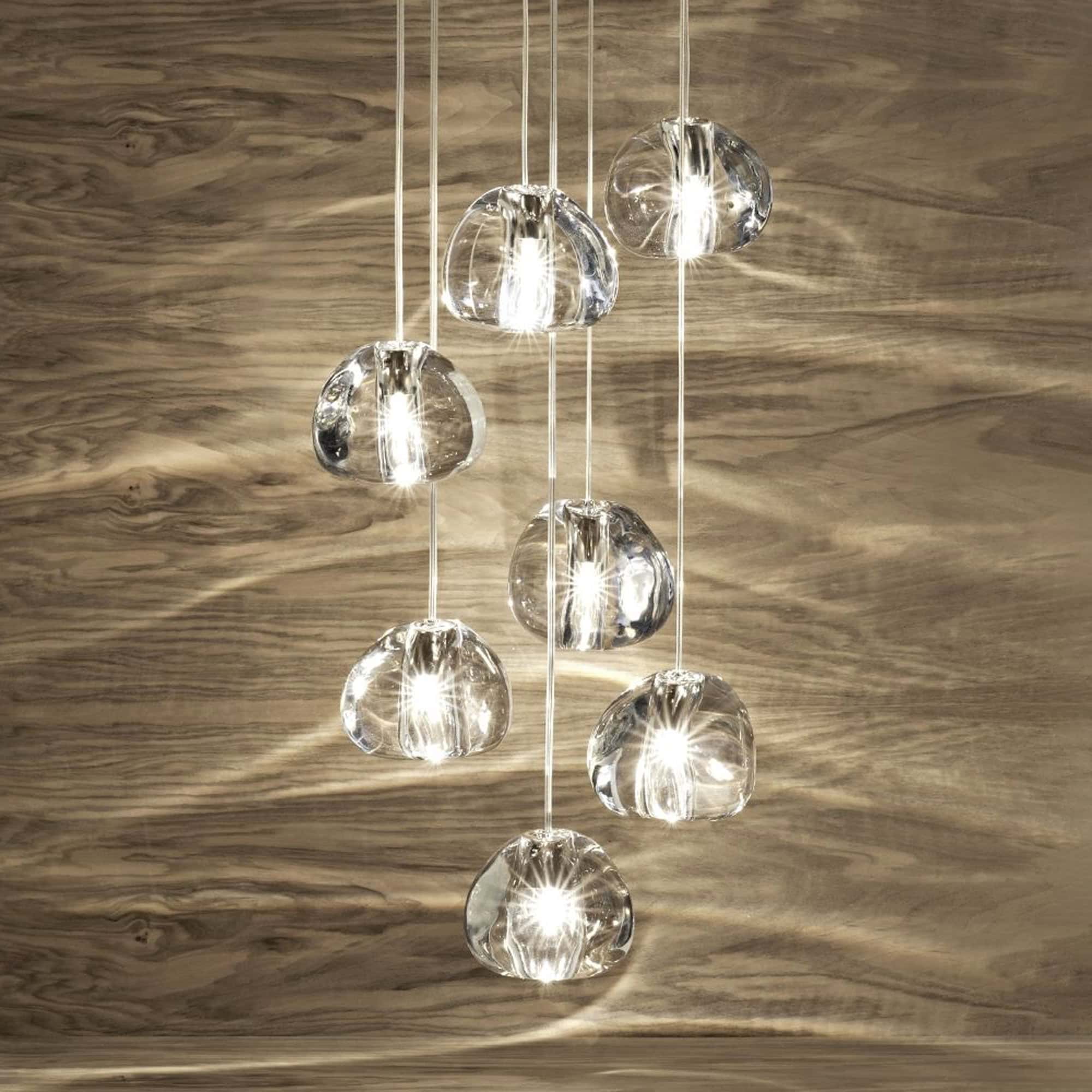 A multi-light pendant fixture with eight clear, irregularly shaped glass globes suspended at various heights against a warm, horizontally grained wooden wall. The lights are illuminated, casting bright reflections.