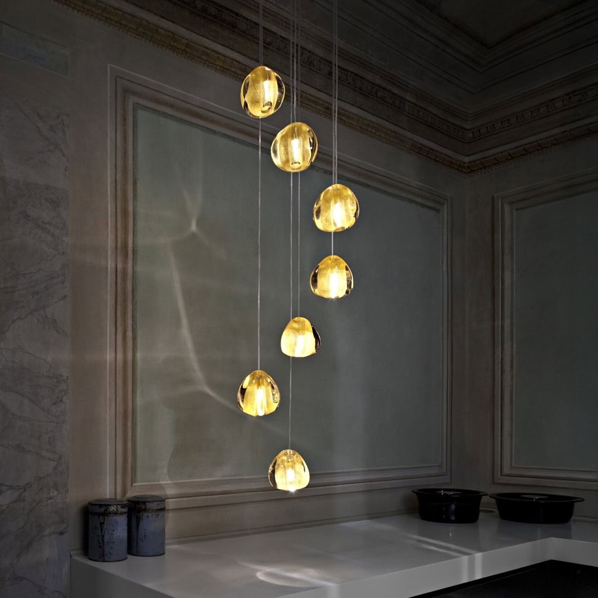 Seven golden, irregularly shaped pendant lights hanging at different heights in a dimly lit, elegant room with classic architectural details and a white countertop.