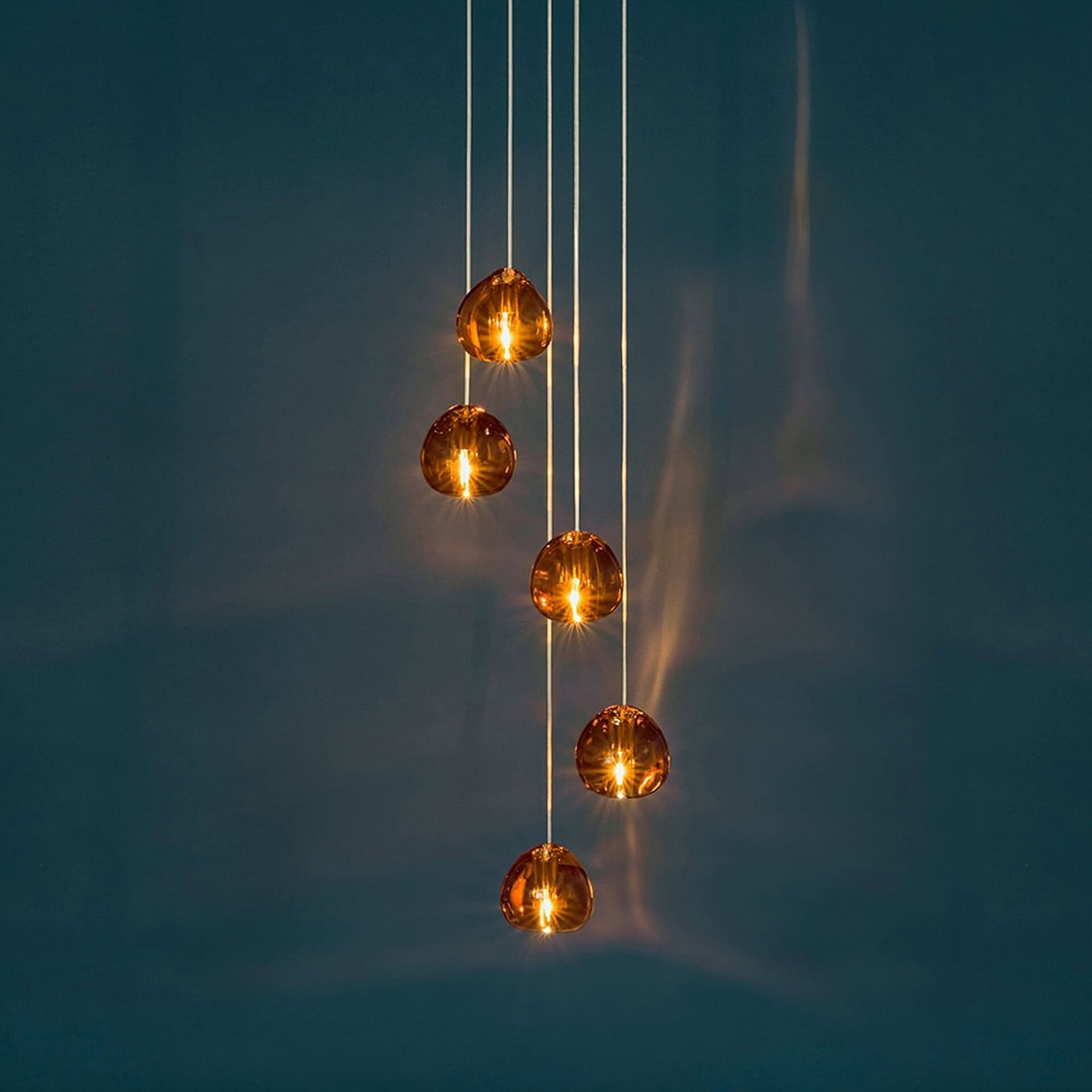 Six illuminated amber glass pendant lights, teardrop-shaped, suspended at varying heights against a dark teal wall.