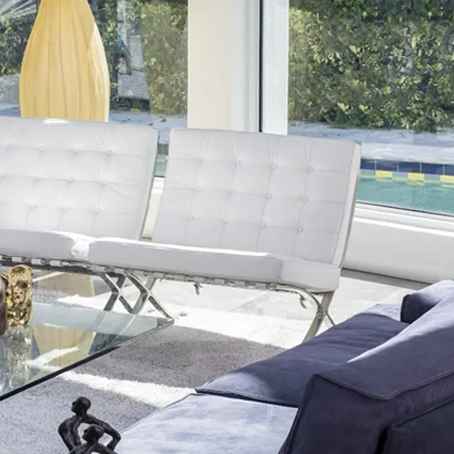 A modern living room with two white tufted chairs, a glass coffee table, and a dark sofa, overlooking a swimming pool and green foliage through large windows.
