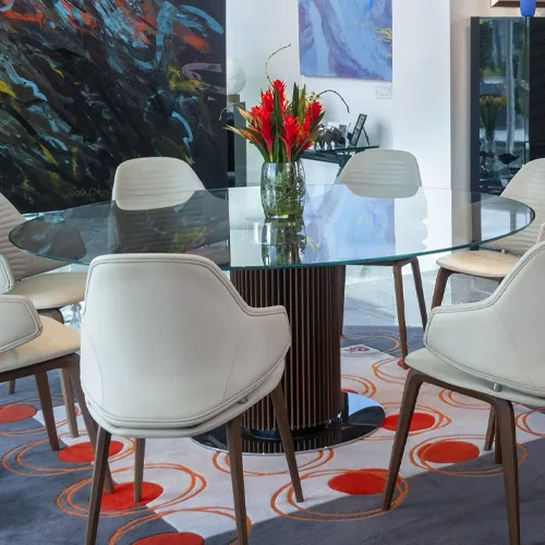 A modern dining room with an oval glass table, cream upholstered chairs, and a vibrant red and yellow floral centerpiece on a patterned grey and orange rug.