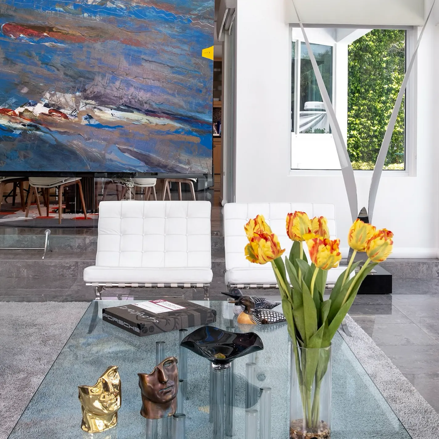 A modern living room with a large abstract painting on the back wall, two white tufted chairs, and a glass coffee table adorned with yellow and red tulips, metallic face sculptures, and a Porsche box. A window showing green foliage is on the right.