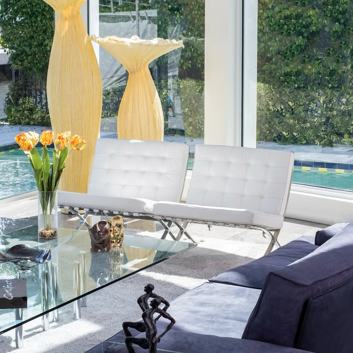 A bright, modern living room with large windows overlooking a pool and waterway. Features a grey sofa, glass coffee table with tulips, white Barcelona chairs, and tall sculptural vases.