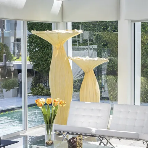 Two large, cream-colored sculptural floor lamps with ruffled tops stand in a modern room with floor-to-ceiling windows overlooking a blue swimming pool and green foliage. A vase of orange and yellow tulips is in the foreground, next to a white tufted sofa.
