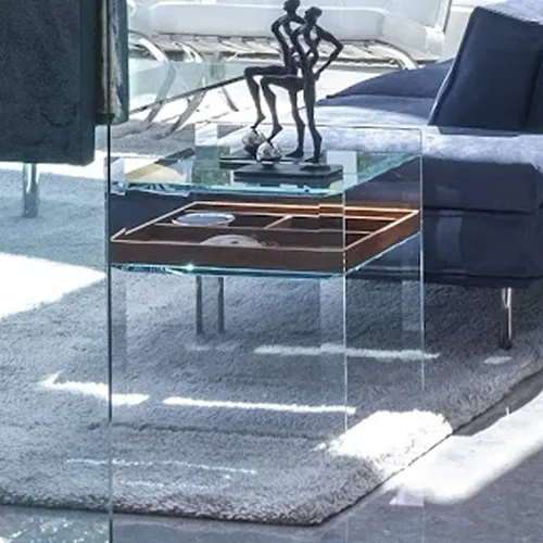 A modern clear glass side table with an abstract dark sculpture and a wooden compartmentalized tray on its shelves, set on a light-colored carpet.