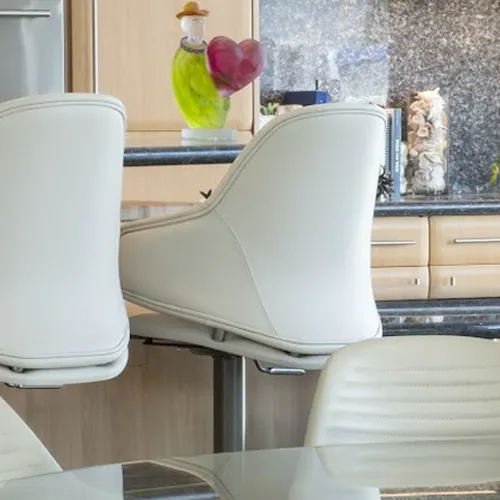 Three cream-colored bar stools at a dark kitchen counter with a green and pink heart figurine, wooden cabinets, and a decorative vase.