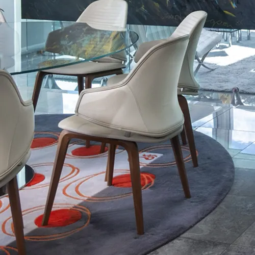 Three modern cream upholstered chairs with wooden legs are arranged around a glass table on a round grey rug with orange and light grey circular patterns. Bright natural light comes from a large window in the background.