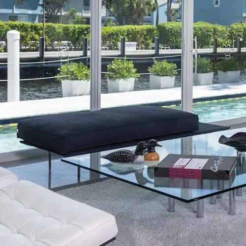 A modern living room with a black daybed, a glass coffee table with duck figurines, and a white tufted sofa, all overlooking a waterfront with white planters and green bushes.