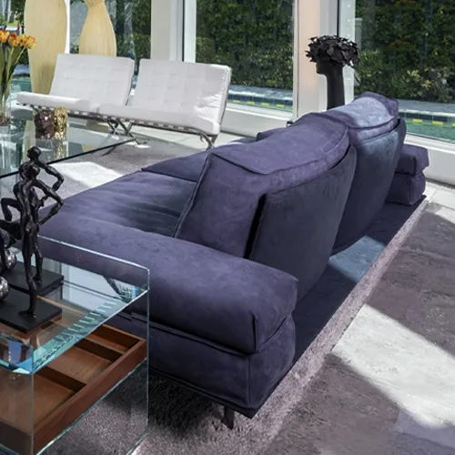 A modern living room with a large indigo sofa, a glass coffee table with abstract art, and white lounge chairs by a large window overlooking greenery.