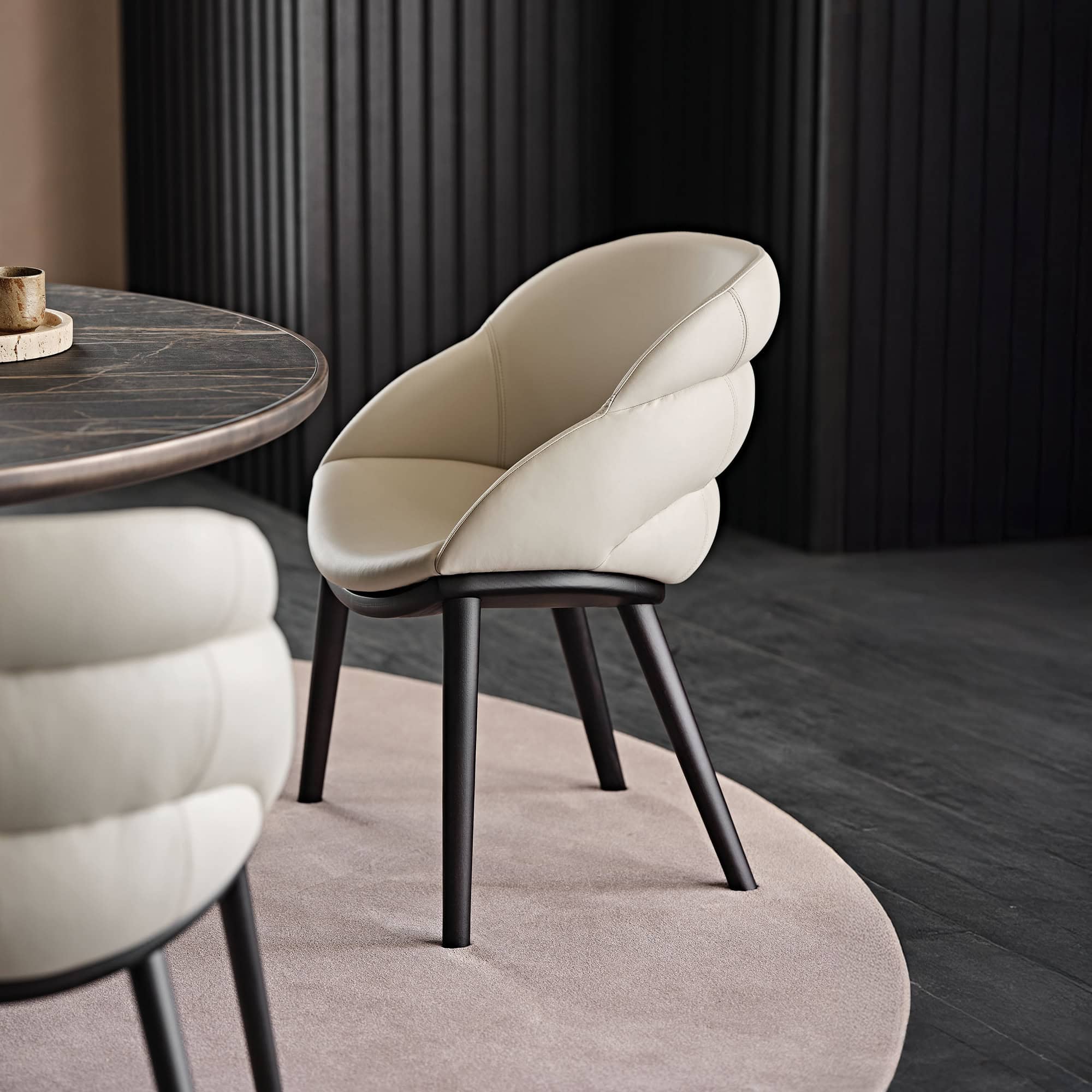 A modern cream upholstered dining chair with dark legs on a round rug, next to a dark marble table in a room with dark fluted wall paneling.