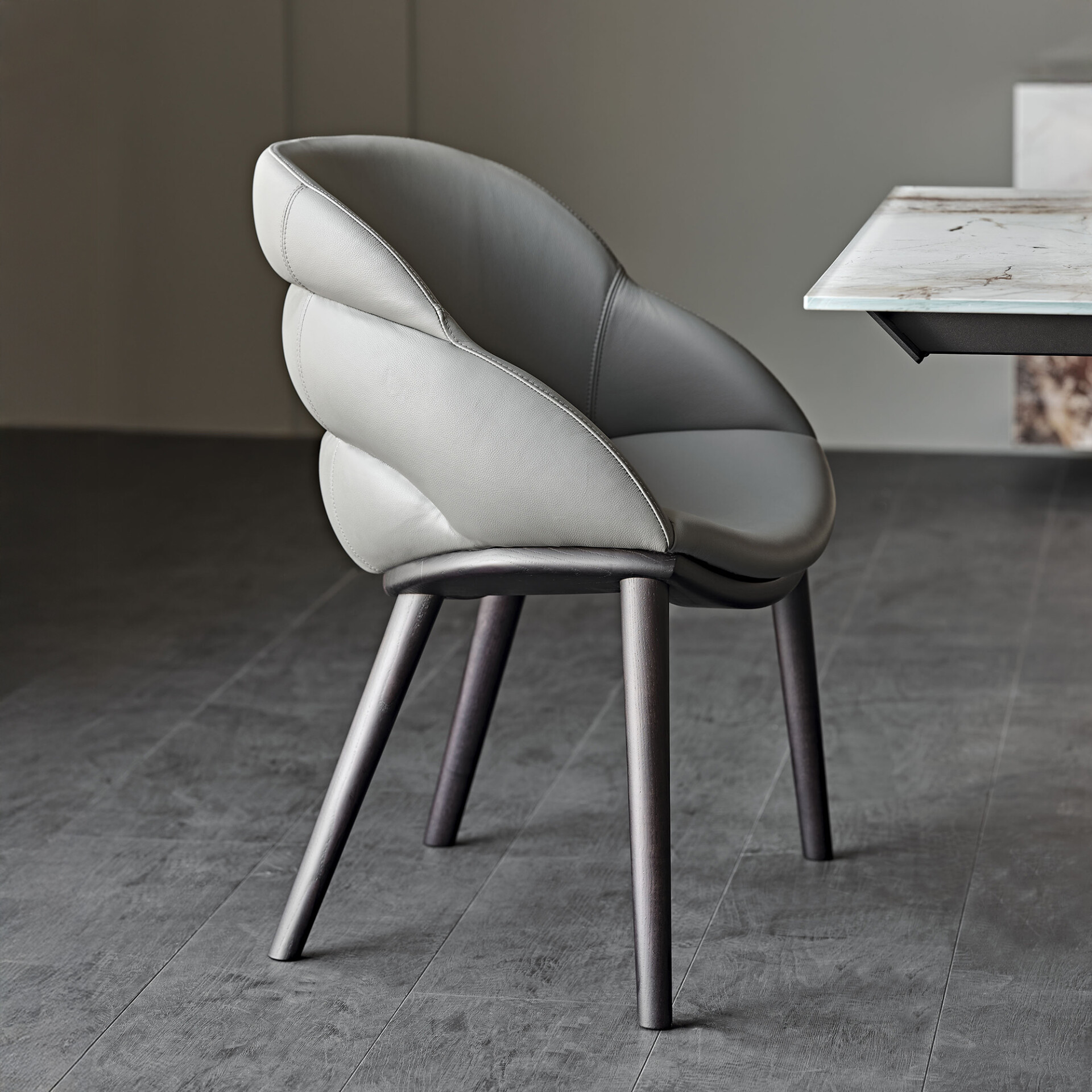 A modern light gray upholstered dining chair with a distinctive folded backrest and dark tapered legs, next to a marble-topped table on a dark gray floor.