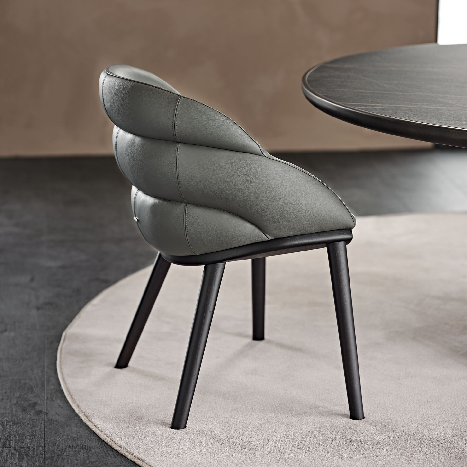 A modern dining chair with a grey ribbed leather back and dark legs, next to a dark wooden table, on a light-colored rug.