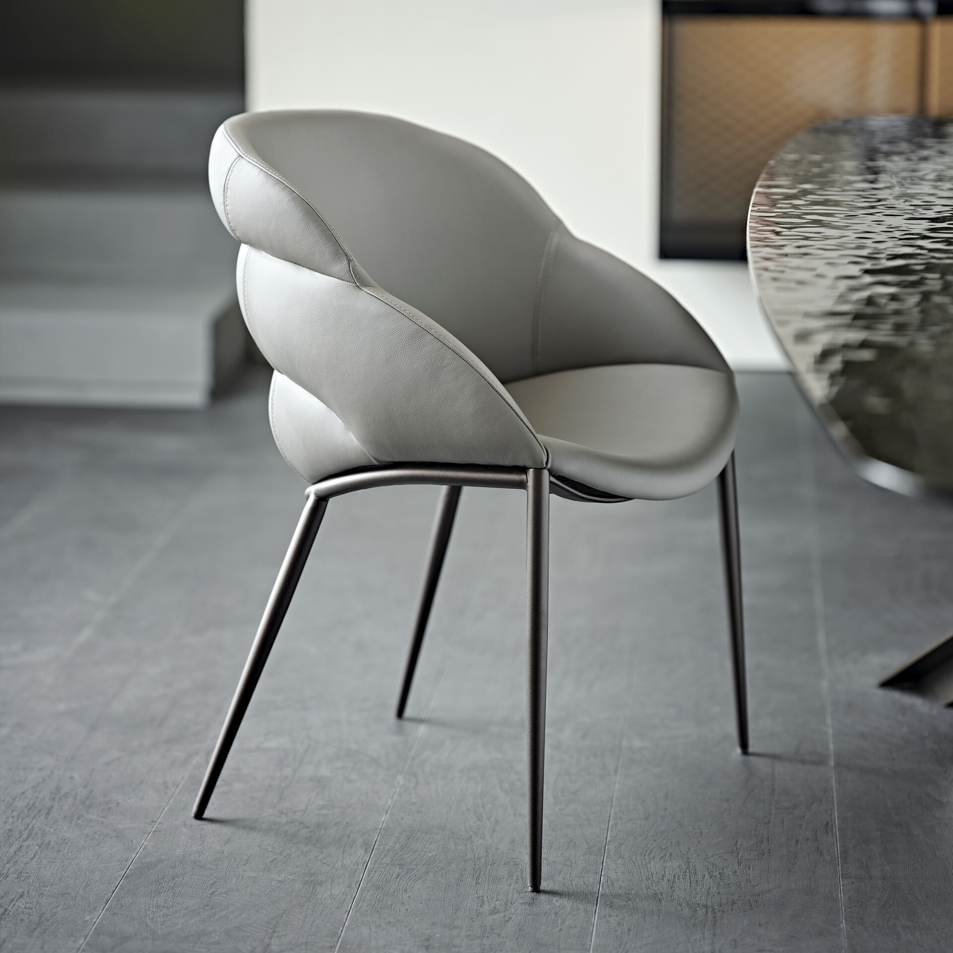A modern light grey upholstered dining chair with a curved back and dark metal legs, positioned on a grey floor next to a dark, textured table.