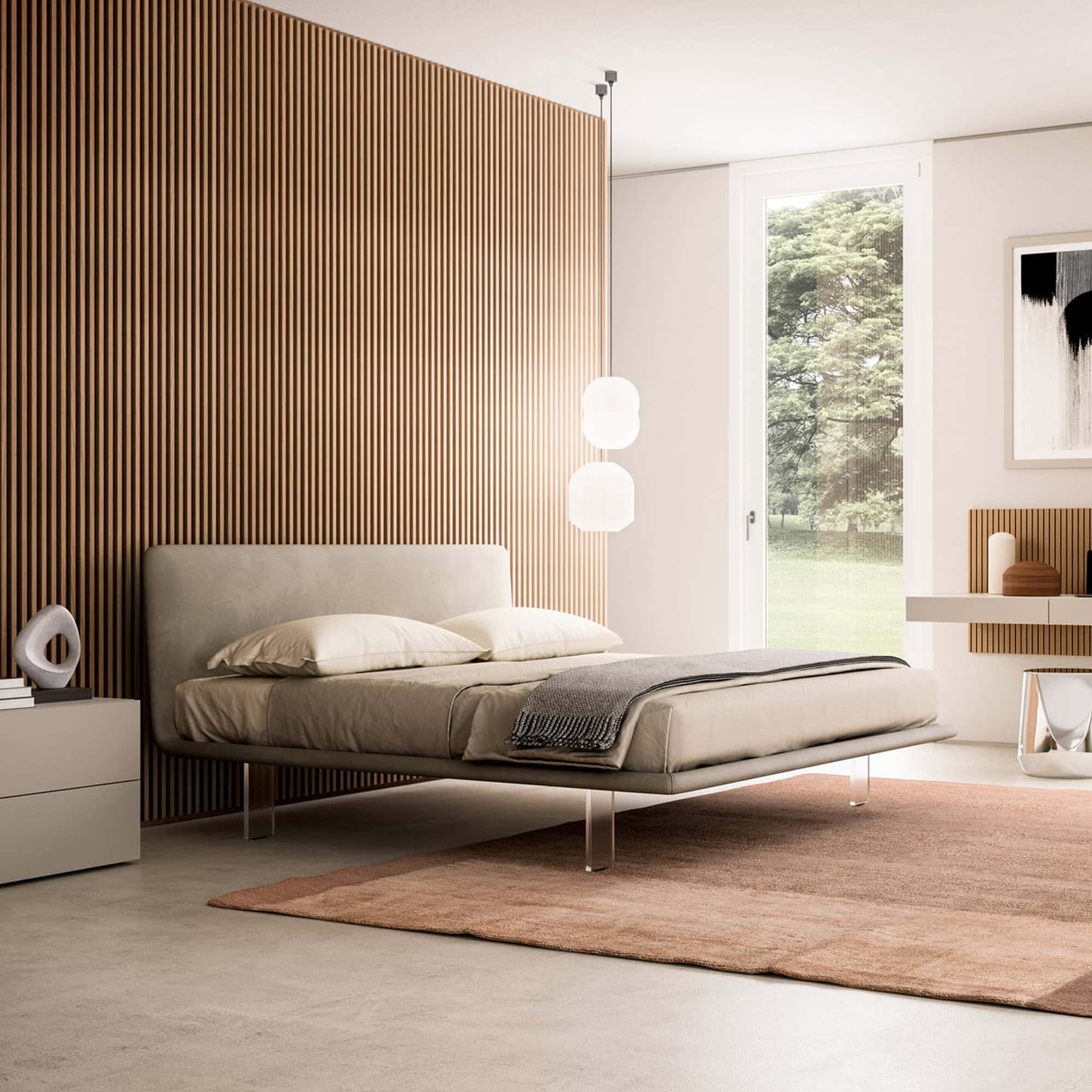 A modern bedroom featuring a low-profile upholstered bed with transparent legs, a slatted wooden accent wall, a large window overlooking green trees, and a terracotta rug.