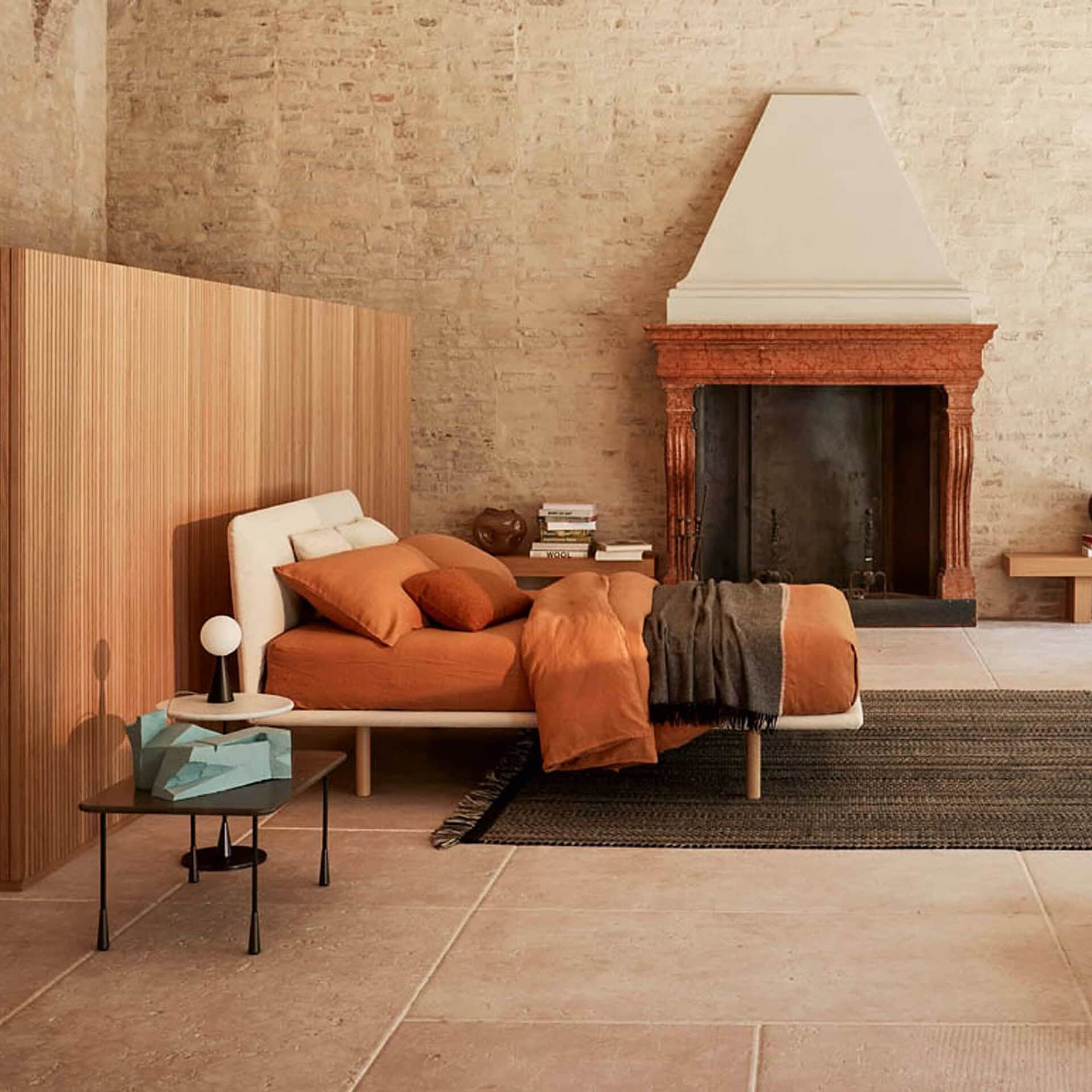 A bedroom with a low bed featuring terracotta bedding, a light wooden slatted wall, and an ornate fireplace against an exposed brick wall. A dark gray rug and stone floor complete the look.