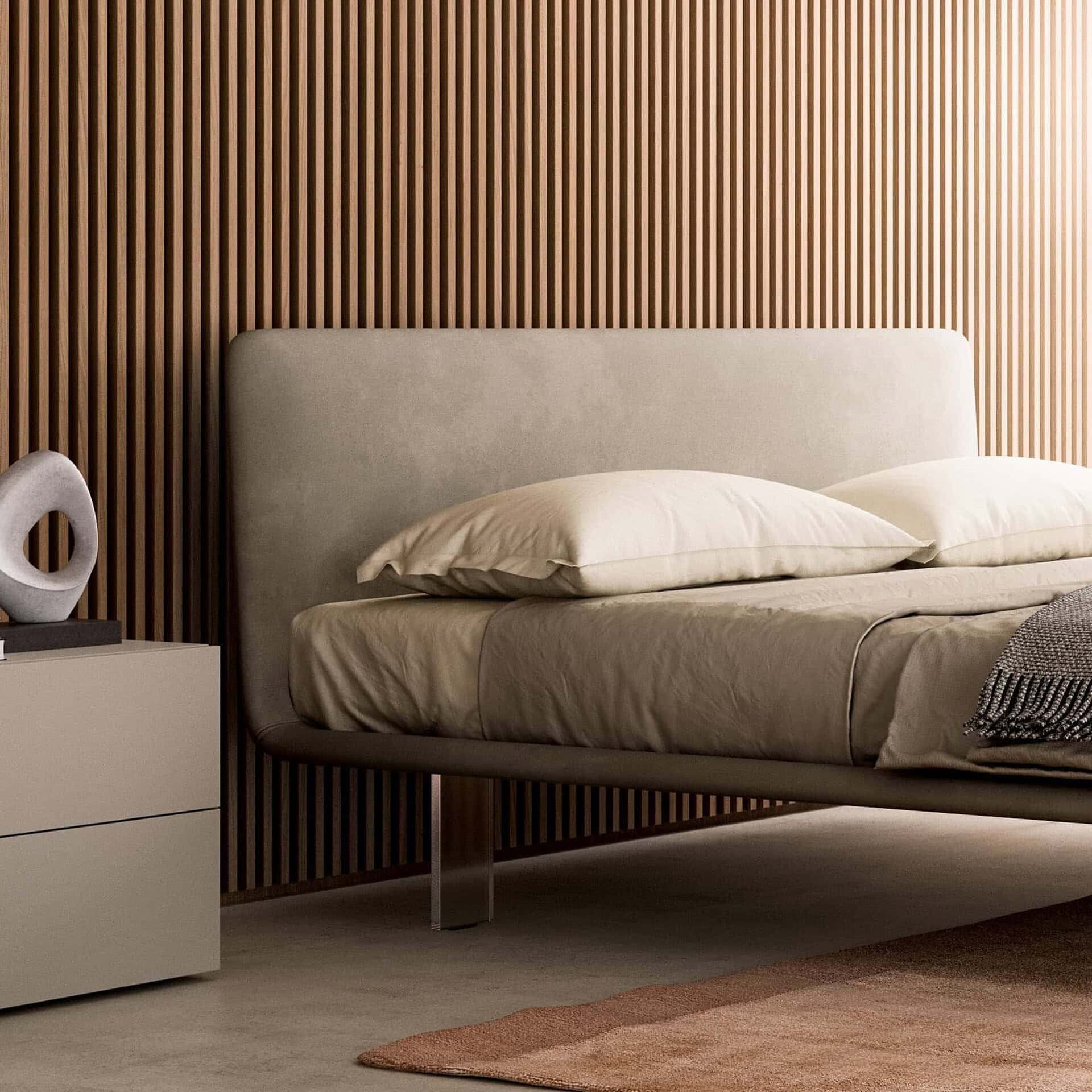 A modern bedroom with a light greige upholstered bed against a vertical wooden slat wall. A matching bedside table with an abstract sculpture is on the left, and a terracotta rug is on the floor.