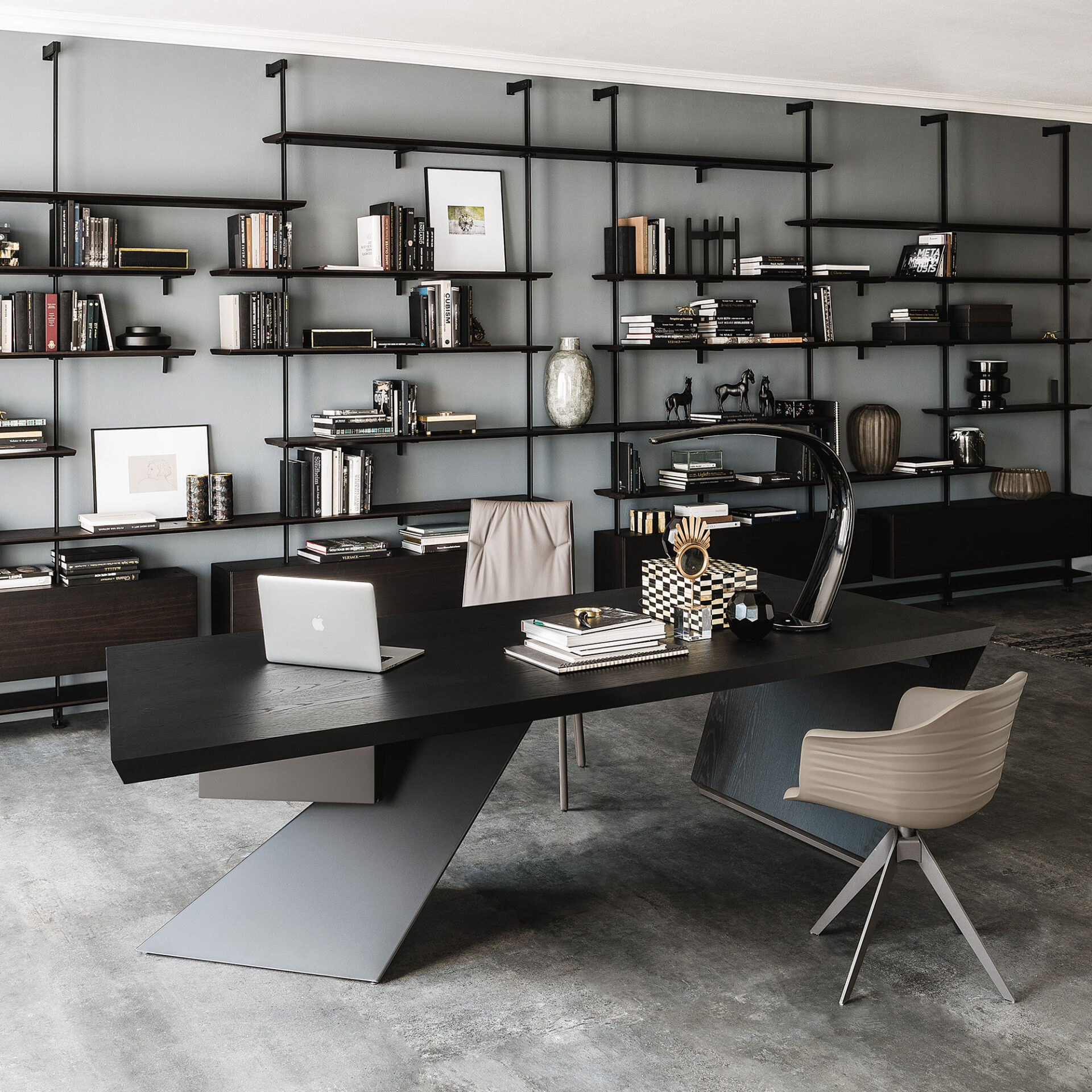 A modern home office with a dark wood desk, angular grey base, and a full-wall black and dark wood shelving unit against a light grey wall. A laptop, desk lamp, and decorative items are on the desk, and the shelves are filled with books and decor. Concrete floor.