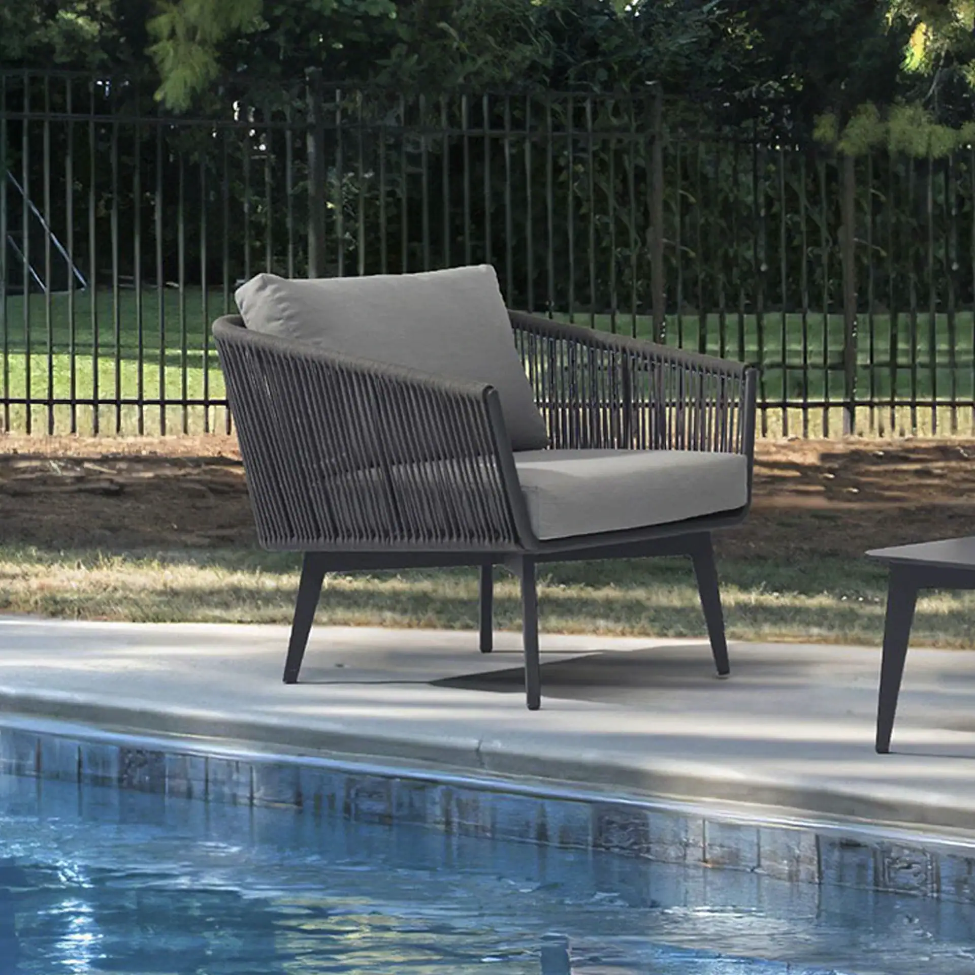 A modern dark gray woven outdoor armchair with light gray cushions sits on a concrete patio next to a blue swimming pool. A dark fence and green trees are in the background.